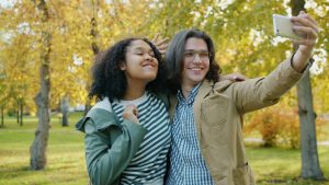 young attractive girl and guy taking selfie in park hugging having fun with hand gestures. Joyful youth and modern lifestyle concept.